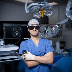 A man with folded arms stands in a dimly lit operating theatre with a controller in his left hand. He wears scrubs, surgical gloves and large dark goggles.