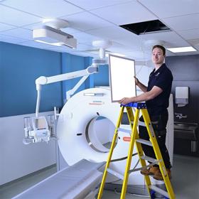 A young man stands on stepladder in a clinical room, alongside a doughnut-shaped scanner, displaying a panel removed from the ceiling above him.